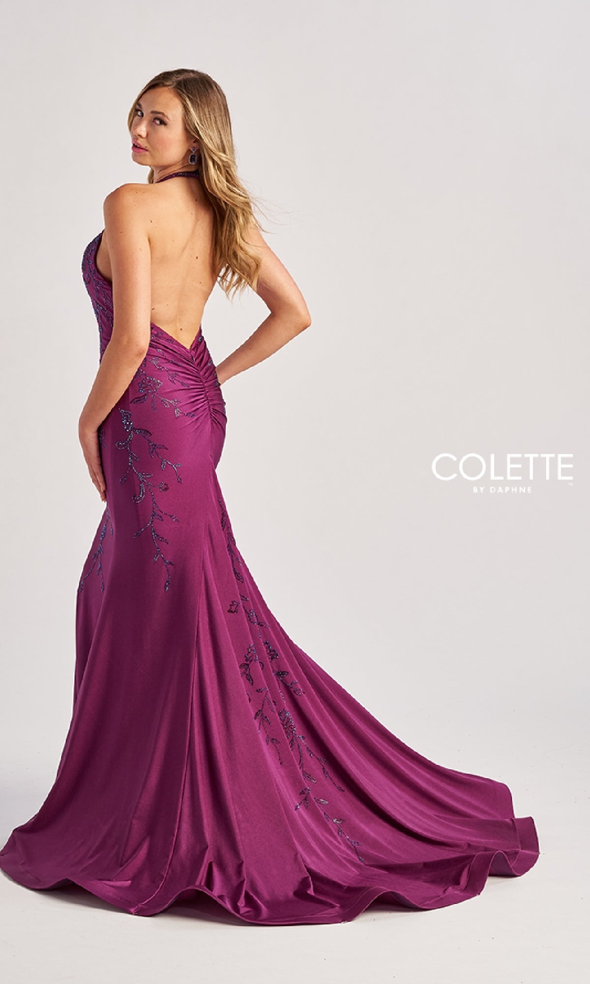 A woman poses in a floor-length magenta mermaid prom dress with beaded floral patterns and a ruched back, branded as Colette CL8455 (Long Halter Prom Dress), against a plain background.
